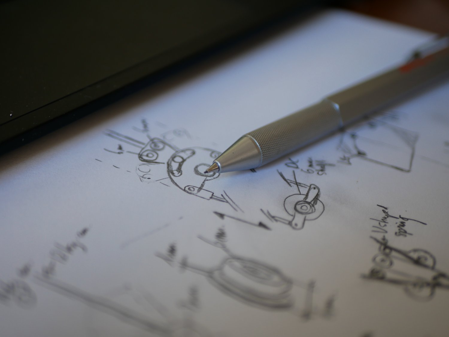 A mechanical pencil mid-sketch over a page of pencil studies of the mechanism: small drawings of cams, levers, springs, with hand-written measurements and notes.