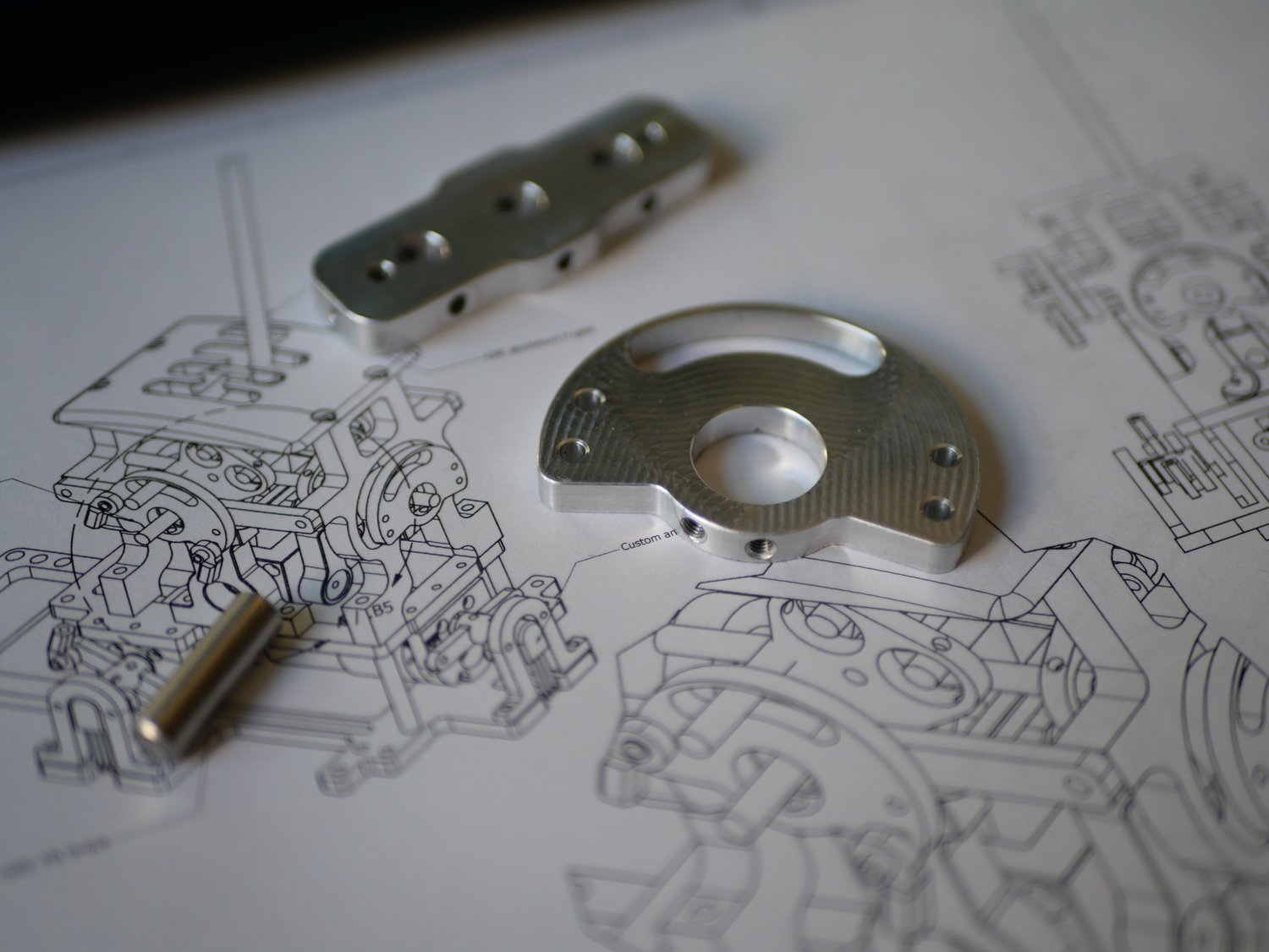 The printed studio sheet with three machined aluminium parts resting on it: a slim bracket, a circular flange with mounting holes, and a small cylindrical pin.