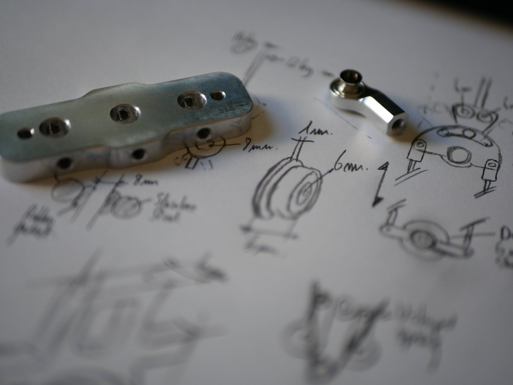 Pencil sketches with measurements alongside a 3D-printed prototype plate and a small machined aluminium linkage.